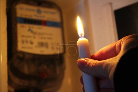 Téléchargez les photos : Bougie à la main brille dans l'obscurité près du compteur d'électricité lors d'une panne de courant à la maison. Ville, pays sans électricité novembre 2022 - en image libre de droit