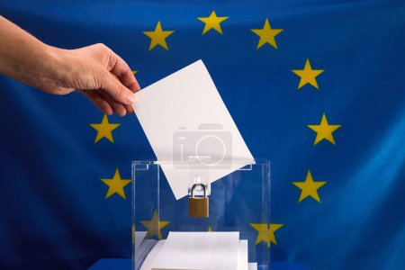 Hand wirft einen Stimmzettel in eine Schachtel mit EU-Flagge im Hintergrund.