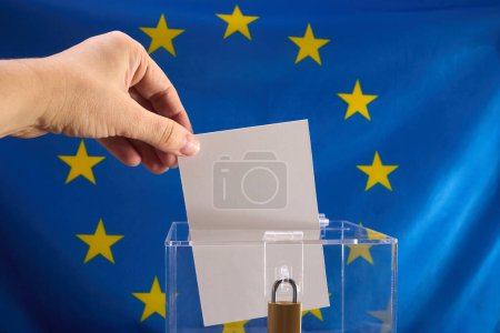 Hand placing a voting ballot into a clear box with EU flag backdrop.