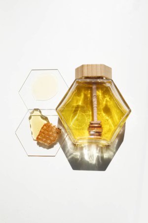 A honey jar with a wooden dipper, honeycomb piece, and drops against a white backdrop.