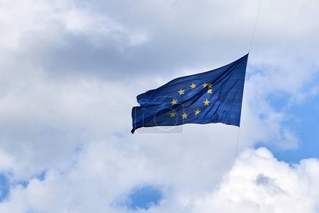 EU flag with circle of stars fluttering against a blue sky with clouds.