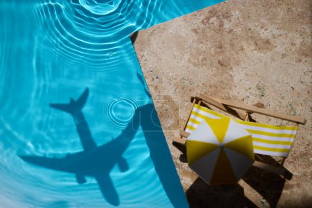 A shadow of a plane over sparkling water next to a deck chair and umbrella.