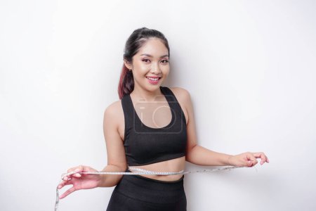 Foto de Retrato de una joven asiática alegre en ropa deportiva midiendo la cintura con cinta adhesiva, sintiéndose feliz por perder peso en casa. Estilo de vida saludable, deportes y adelgazamiento concepto de dieta - Imagen libre de derechos