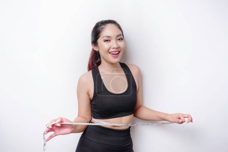 Foto de Retrato de una joven asiática alegre en ropa deportiva midiendo la cintura con cinta adhesiva, sintiéndose feliz por perder peso en casa. Estilo de vida saludable, deportes y adelgazamiento concepto de dieta - Imagen libre de derechos
