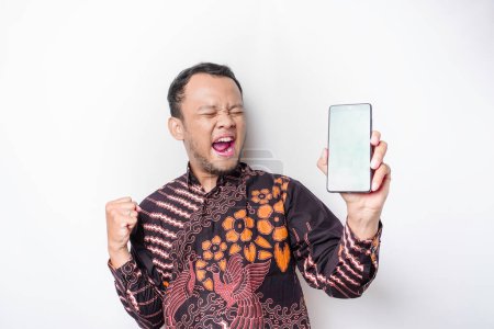 Photo for A happy young Asian man wearing batik shirt showing successful expression showing copy space on his phone isolated by white background - Royalty Free Image