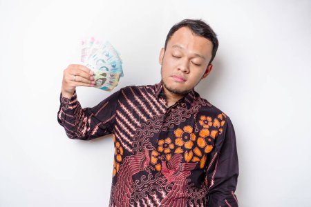 Foto de Joven asiático lleva camisa batik sosteniendo ventilador de dinero en efectivo en indonesio rupiah billetes haciendo aislado en fondo blanco estudio retrato - Imagen libre de derechos