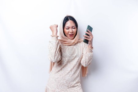 Photo for A young Asian Muslim woman with a happy successful expression wearing a hijab and holding smartphone isolated by white background - Royalty Free Image