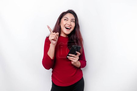 Téléchargez les photos : Femme asiatique excitée portant haut rouge pointant vers l'espace de copie sur elle tout en tenant son téléphone, isolé par fond blanc - en image libre de droit