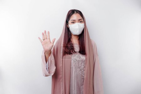 Foto de Joven mujer musulmana asiática usando una máscara y hiyab muestra su gesto de palma o stop, concepto de prevención covid - Imagen libre de derechos