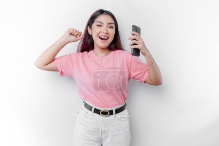 Téléchargez les photos : Une jeune femme asiatique avec une expression heureuse et réussie portant un t-shirt rose et tenant un smartphone isolé par un fond blanc - en image libre de droit