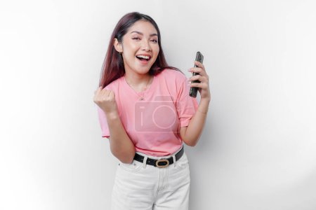 Photo for A young Asian woman with a happy successful expression wearing pink t-shirt and holding smartphone isolated by white background - Royalty Free Image