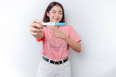 Téléchargez les photos : Happy consciente reconnaissante jeune femme portant un t-shirt rose montrant son test de grossesse, la main sur la poitrine souriant isolé sur fond blanc - en image libre de droit