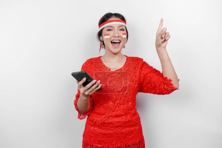 Téléchargez les photos : Une femme asiatique heureuse portant kebaya rouge et bandeau, tenant son téléphone, et pointant l'espace de copie sur elle, isolée par un fond blanc. Jour de l'indépendance de l'Indonésie - en image libre de droit