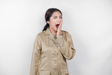 Téléchargez les photos : Portrait de femme asiatique endormie portant un uniforme marron et couvrant sa bouche d'une main en bâillant. Image isolée sur fond blanc - en image libre de droit