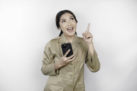 Photo for Smiling government worker woman holding her smartphone and pointing to copy space above her. PNS wearing khaki uniform. - Royalty Free Image