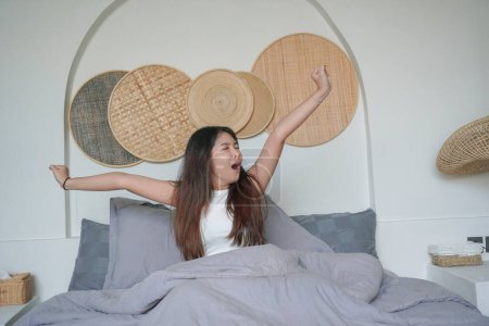 Sleepy Asian young woman yawning and raising hand waking up in early morning after deep sleep, peaceful morning holiday concept