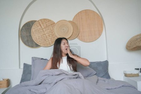 Sleepy Asian young woman yawning and raising hand waking up in early morning after deep sleep, peaceful morning holiday concept