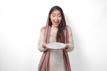 Surprised Asian Muslim woman in headscarf veil hijab holding plate with copy space inside, isolated white background
