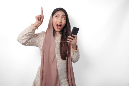Surprised Asian Muslim woman in veil hijab holding a smart phone while pointing up to the copy space upwards over isolated white background. Ramadan concept