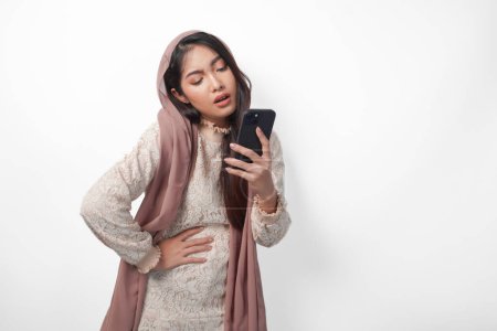 Joven mujer musulmana asiática en el hiyab velo sintiendo hambre mientras sostiene su estómago y el uso de un teléfono inteligente para ordenar alimentos. Concepto Ramadán y Eid Mubarak