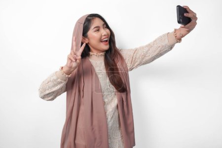 Hermosa mujer musulmana asiática en velo hijab pañuelo riendo y tomando un selfie divertido usando el teléfono móvil, aislado por el fondo blanco