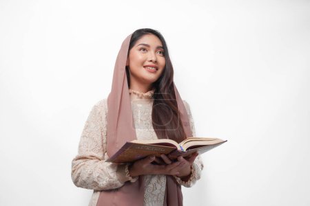 Serious Asian Muslim woman wearing veil hijab praying and reading Al Quran, standing over isolated white background. Ramadan and Eid Mubarak concept