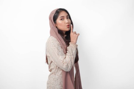 Young Asian Muslim woman wearing headscarf veil hijab looking to camera while putting a finger in front of lips gesturing stay silent or stay quiet, isolated by white background