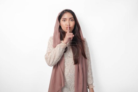 Junge asiatische Muslimin mit Kopftuchschleier-Hijab blickt in die Kamera, während sie gestikulierend einen Finger vor die Lippen legt, schweigt oder schweigt, isoliert von weißem Hintergrund
