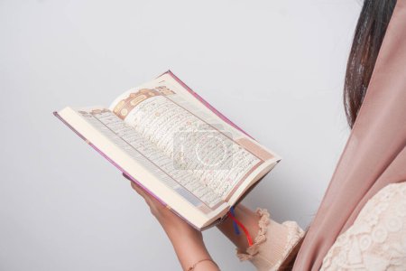 Close up portrait of Asian Muslim woman in headscarf veil hijab holding and reading Al Quran over isolated white background. Ramadan and Eid Mubarak concept.