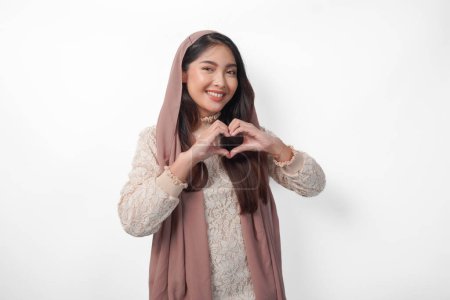 A happy young Asian Muslim woman wearing veil hijab making love heart shape using hands on isolated white background. Ramadan and Eid Mubarak concept