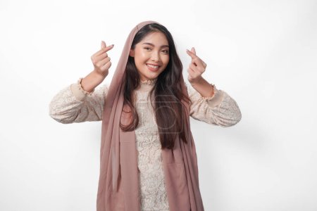 A happy young Asian Muslim woman wearing veil hijab making love heart shape using hands on isolated white background. Ramadan and Eid Mubarak concept