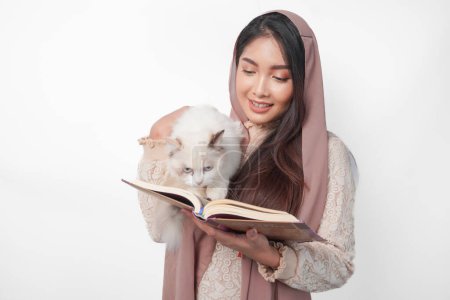 Attraktive junge asiatische Muslimin im verschleierten Hijab lächelt, während sie ein weißes Haustier mit Stoffpuppen umarmt und auf der anderen Seite den Al Quran in der Hand hält