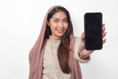 Lächelnde junge asiatische Muslimin im Kopftuchschleier-Hijab präsentiert oder zeigt Kopierraum auf dem Smartphone-Bildschirm, isoliert von einem Studio mit weißem Hintergrund. Ramadan und Eid Mubarak-Konzept.