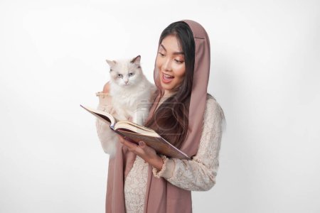 Attraktive junge asiatische Muslimin im verschleierten Hijab lächelt, während sie ein weißes Haustier mit Stoffpuppen umarmt und auf der anderen Seite den Al Quran in der Hand hält