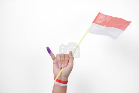 Group of hand wearing flag ribbon on wrist showing little finger dipped in purple ink after voting for Indonesia Election or Pemilu while holding mini flag, isolated over white background