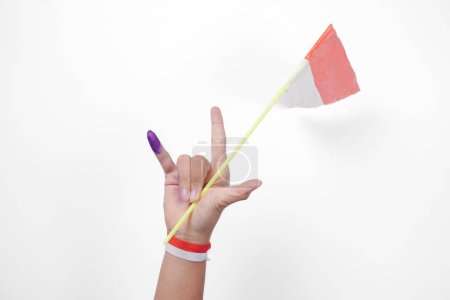 Group of hand wearing flag ribbon on wrist showing little finger dipped in purple ink after voting for Indonesia Election or Pemilu while holding mini flag, isolated over white background