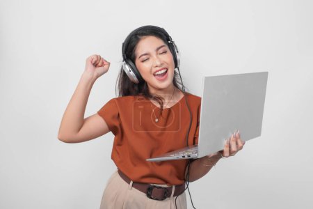 Photo for Young woman wearing brown shirt and headphone while holding laptop to listen music with clenched fist gesture isolated over white background. - Royalty Free Image