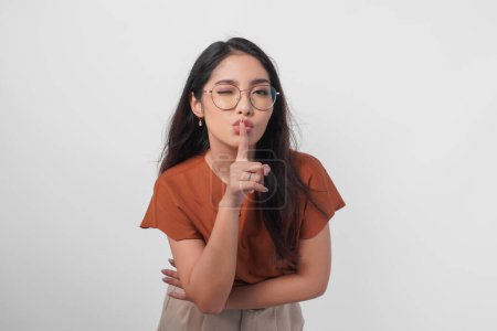 Young Asian woman wearing brown shirt and eyeglass looking to camera while putting a finger in front of lips gesturing stay silent or stay quiet, isolated by white background.