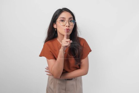 Young Asian woman wearing brown shirt and eyeglass looking to camera while putting a finger in front of lips gesturing stay silent or stay quiet, isolated by white background.