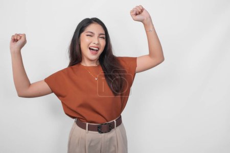 Photo for A young Asian woman in brown shirt with a happy successful expression and raising up clenched fist in excitement isolated by white background. - Royalty Free Image