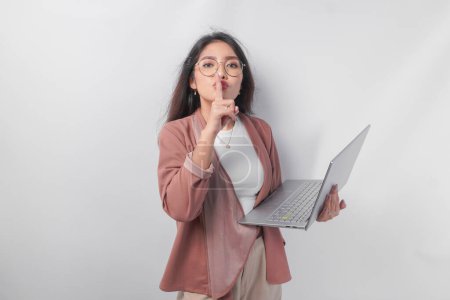 Junge asiatische Geschäftsfrau, die gestikulierend einen Finger vor die Lippen legt, schweigt oder schweigt, während sie ihren Laptop hält, isoliert von weißem Hintergrund.