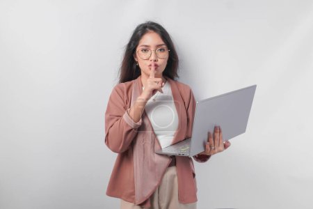 Junge asiatische Geschäftsfrau, die gestikulierend einen Finger vor die Lippen legt, schweigt oder schweigt, während sie ihren Laptop hält, isoliert von weißem Hintergrund.