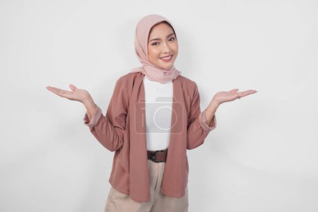 Lächelnde junge asiatische Muslimin im Hijab präsentiert Kopierraum auf ihrer rechten und linken Seite vor isoliertem weißem Hintergrund.