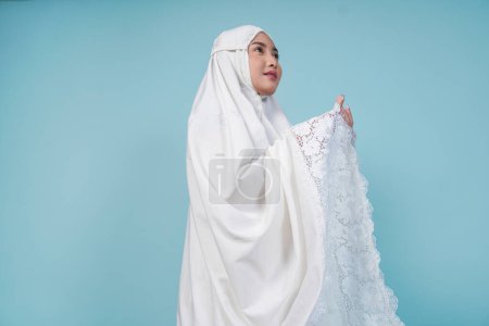 Gros plan portrait de la jeune femme musulmane asiatique portant une robe de prière posant avec les mains, isolée par un fond bleu. Concept du Hadj.