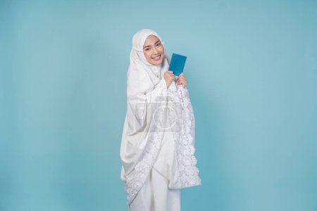 Happy young Asian Muslim woman wearing prayer gown holding passport isolated by blue background. Hajj concept.