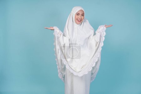 Jeune femme musulmane asiatique souriante portant une robe de prière présentant un espace de copie à côté d'elle, isolée par un fond bleu. Concept du Hadj.