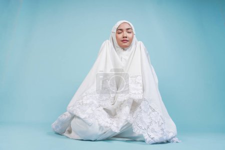 Jeune femme musulmane asiatique en robe de prière assise et priant les yeux fermés sur un fond bleu isolé. Concept du Hadj.