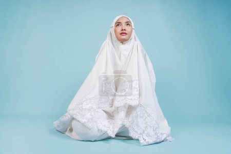 Jeune femme musulmane asiatique en robe de prière assise et priant solennellement les yeux ouverts sur un fond bleu isolé. Concept du Hadj.