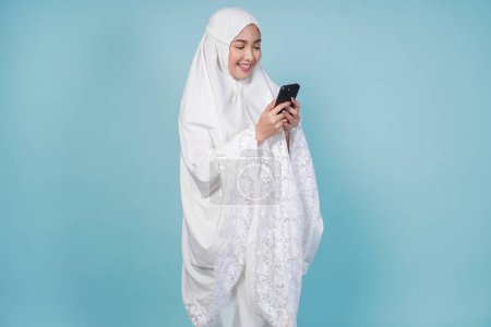 Jeune femme musulmane asiatique en robe de prière en utilisant le téléphone pour envoyer des SMS ou naviguer sur Internet, isolé par fond bleu. Concept du Hadj.