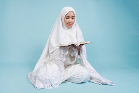 Sérieuse jeune femme musulmane asiatique en robe de prière assise et lisant le Coran sur fond bleu isolé. Concept du Hadj.